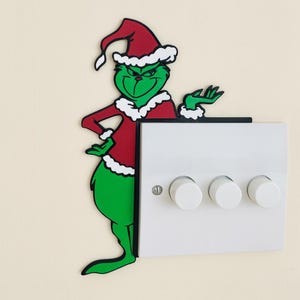 May include: A green cartoon character in a red and white Santa hat and jacket, next to a white switch plate with three white dimmer switches. The character is leaning against the switch plate.