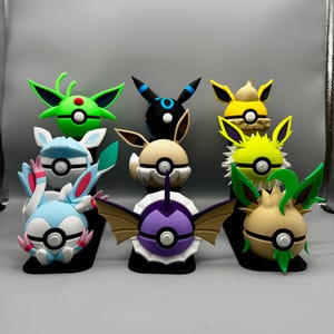 May include: A collection of colorful Pokémon character figures, each designed with unique features. The figures are displayed on black bases, showcasing a variety of colors including green, black, yellow, blue, and purple. The figures are round with detailed, stylized features.