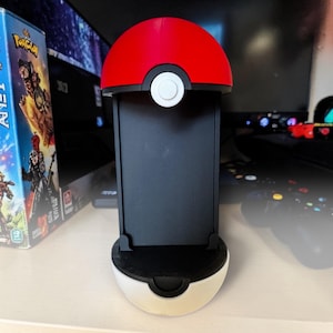 Puede incluir: Un soporte vertical para consola de juegos dise&ntilde;ado para parecerse a una Pok&eacute; Ball roja y blanca. El soporte presenta una semiesfera superior roja brillante, una banda central negra con un bot&oacute;n circular blanco y una base negra.
