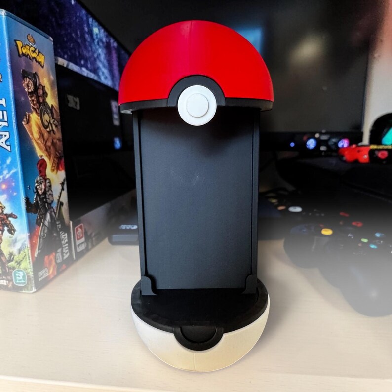 Puede incluir: Un soporte vertical para consola de juegos dise&ntilde;ado para parecerse a una Pok&eacute; Ball roja y blanca. El soporte presenta una semiesfera superior roja brillante, una banda central negra con un bot&oacute;n circular blanco y una base negra.