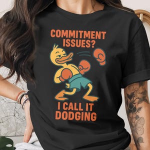May include: Black t-shirt featuring a cartoon duck wearing boxing gloves and shorts, dodging punches. Text reads "COMMITMENT ISSUES? I CALL IT DODGING" in orange and white. The design is centered on the shirt.