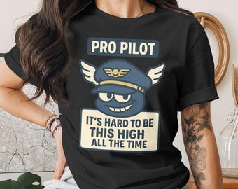 May include: Black t-shirt with a humorous graphic. The design features a cartoon pilot's hat with wings and a smiling face. Text reads "PRO PILOT" above and "IT'S HARD TO BE THIS HIGH ALL THE TIME" below the graphic.