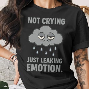 May include: Black t-shirt with the text "NOT CRYING JUST LEAKING EMOTION." A gray cloud with a sad face and rain drops is printed on the front. The shirt is a casual wear item.