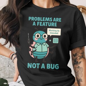 May include: Black t-shirt with the text "PROBLEMS ARE A FEATURE NOT A BUG". The shirt features a cartoon bug character with glasses, holding a screwdriver and a small device. The bug has a speech bubble that says "Working As Intended".