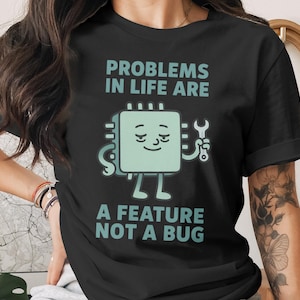 May include: Black t-shirt with the text "PROBLEMS IN LIFE ARE A FEATURE NOT A BUG" in teal. The graphic is a smiling computer chip holding a wrench. The shirt is a short-sleeved crew neck.