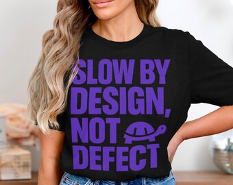 May include: Black t-shirt with the text "SLOW BY DESIGN, NOT DEFECT" in purple block letters. A purple turtle graphic is included. The shirt is a crew neck and appears to be made of cotton.