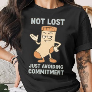 Puede incluir: Camiseta negra con el texto "NOT LOST" sobre un personaje de calcetín de dibujos animados. El calcetín tiene una expresión divertida y el texto "JUST AVOIDING COMMITMENT" debajo. El calcetín es beige y naranja.