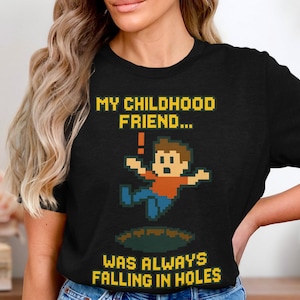 May include: Black t-shirt with a pixelated graphic of a person falling into a hole. The text reads "MY CHILDHOOD FRIEND... WAS ALWAYS FALLING IN HOLES" in yellow pixelated font. The shirt is made of a soft material.