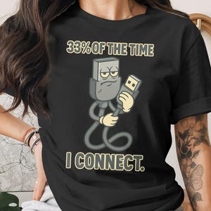 May include: Black t-shirt featuring a cartoon USB plug character holding a USB port. The text reads "33% OF THE TIME I CONNECT." The design is in shades of gray, yellow, and black.