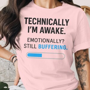 May include: Light pink t-shirt with the text "TECHNICALLY I'M AWAKE. EMOTIONALLY? STILL BUFFERING." in black and blue. A blue progress bar graphic is below the text. The shirt is a casual wear item.