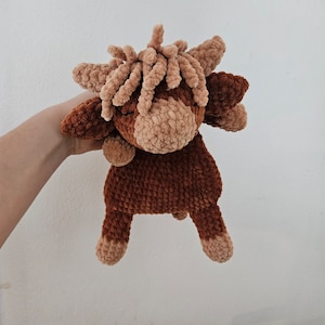 May include: A brown and tan crocheted stuffed animal shaped like a highland cow. The cow has a long, curly mane and a small, brown nose.