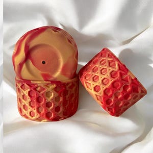 May include: Two decorative, red and yellow geometric patterned containers. One is cylindrical with a lid, and the other is a square shape. The containers have a marbled effect with a mix of red and yellow colors.