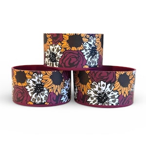 May include: Three cylindrical silicone boots for drinkware, featuring a repeating pattern of sunflowers, dark red roses, and white flowers with black centers. The base of each boot is a solid deep maroon color, providing a stable, protective cover.