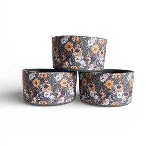 May include: Three cylindrical pet bowls with a dark olive green base color. The exterior surface features a repeating pattern of German Shepherd dog faces interspersed with yellow sunflowers and small white daisy-like flowers throughout.