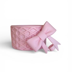 May include: A light pink silicone bracelet featuring a raised geometric lattice pattern across the band. Attached to the side of the band is a prominent three-dimensional bow element with two trailing ribbon ends and a small circular loop component.