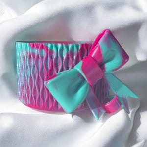 May include: A decorative container with a matching bow, featuring a vibrant swirl of pink and teal colors. The container has a textured, ridged surface. The bow is attached to the container, creating a cohesive design.