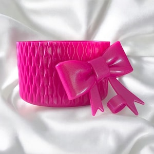 May include: A bright pink, cylindrical container with a textured, wave-like pattern. A matching pink bow is attached to the side. The items are set against a white, silky background.