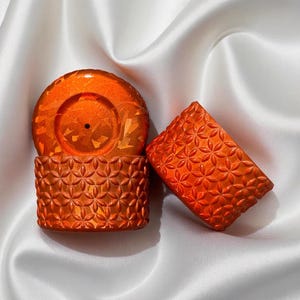 May include: Orange decorative objects with a floral pattern. The objects are arranged on a white satin surface. The objects include a cylinder, a lid, and a rectangular piece.