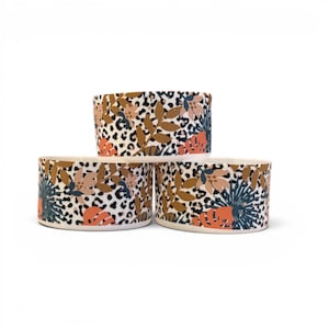 May include: Three cylindrical ceramic bowls stacked in a pyramid formation. Each bowl features a white background with a black leopard print pattern overlaid with colorful illustrations of tropical leaves and flowers in shades of orange, brown, and blue.
