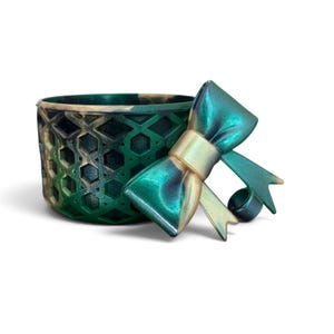 May include: A cylindrical container featuring a dark green and gold marbled finish with a geometric honeycomb pattern on the exterior. A matching decorative bow with a metallic sheen is attached to the side of the container, adding dimension.