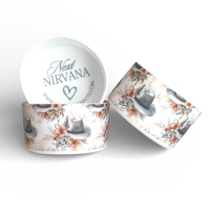May include: Two white ceramic bowls featuring a repeating pattern of gray cowboy hats and orange flowers. The interior of one bowl displays the text Next NIRVANA with a heart symbol and a website URL printed in dark gray on the bottom surface.
