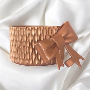 May include: A bronze-colored, cylindrical container with a textured, wave-like pattern. A matching bronze bow is attached to the side. The items are set against a white, silky background.