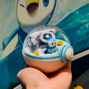 May include: A snow globe with a blue and white cartoon character inside a clear, spherical enclosure. The base is white and blue, with a light blue and white striped knob. The background shows a matching cartoon character on a computer monitor.