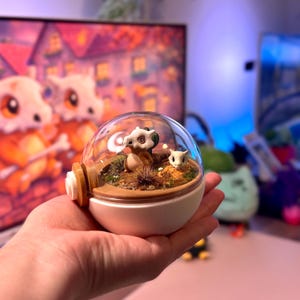 Puede incluir: Un terrario esférico transparente con una base blanca y un acento marrón. En el interior, se exhiben dos pequeñas figuras de dibujos animados de un Pokémon Cubone en medio de plantas y tierra en miniatura. El terrario se sostiene en la mano de una persona.