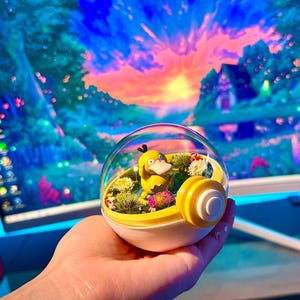 Puede incluir: Un terrario amarillo y blanco en forma de bola, con un personaje de dibujos animados amarillo, plantas verdes y flores rosas. El terrario se sostiene en una mano, con un paisaje colorido visible en el fondo.