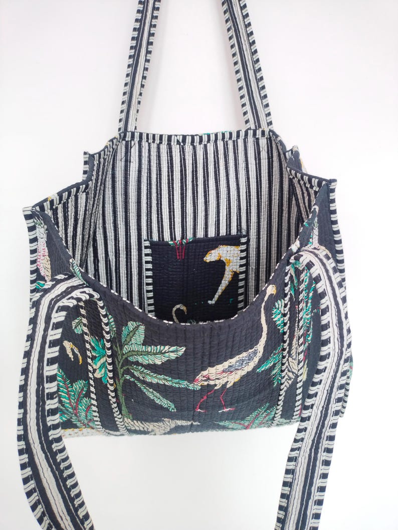 K&ouml;nnte beinhalten: Eine schwarze und wei&szlig;e gestreifte Tragetasche mit einem Blumendruck auf dem Boden. Die Tasche hat eine gro&szlig;e Tasche im Inneren und zwei lange Riemen.