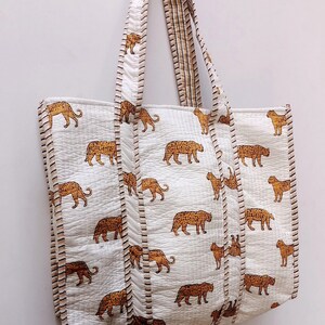 May include: White tote bag featuring a repeating pattern of brown leopards. The bag has matching handles and trim with a striped design. The bag is made of quilted fabric.