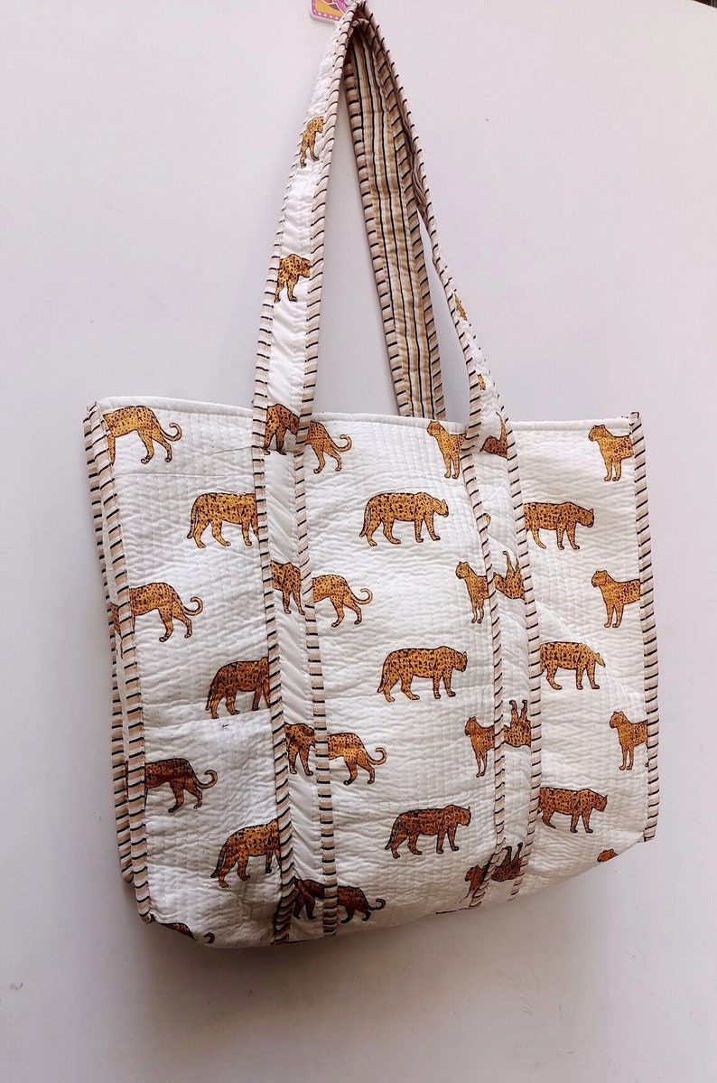 May include: White tote bag featuring a repeating pattern of brown leopards. The bag has matching handles and trim with a striped design. The bag is made of quilted fabric.
