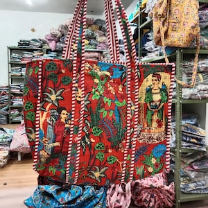Könnte beinhalten: Eine große rote Tragetasche mit weißen und roten Streifen an den Henkeln. Die Tasche ist aus Stoff und hat ein Blumenmuster mit einem Frauenkopf in der Mitte. Die Tasche liegt auf einem Stapel anderer Stofftaschen.