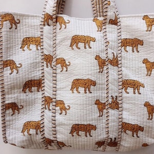 May include: A white tote bag featuring a repeating pattern of brown leopards. The bag has a quilted texture and is trimmed with brown stitching. The handles are white and quilted.