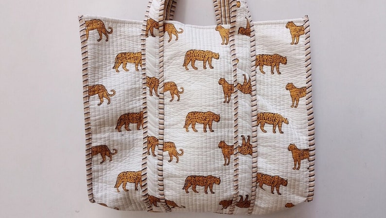 May include: A white tote bag featuring a repeating pattern of brown leopards. The bag has a quilted texture and is trimmed with brown stitching. The handles are white and quilted.
