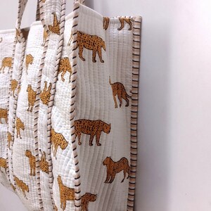 May include: White quilted tote bag featuring a repeating pattern of brown leopard illustrations. The bag has matching handles and trim with a striped design. The bag is made of a soft, quilted material.