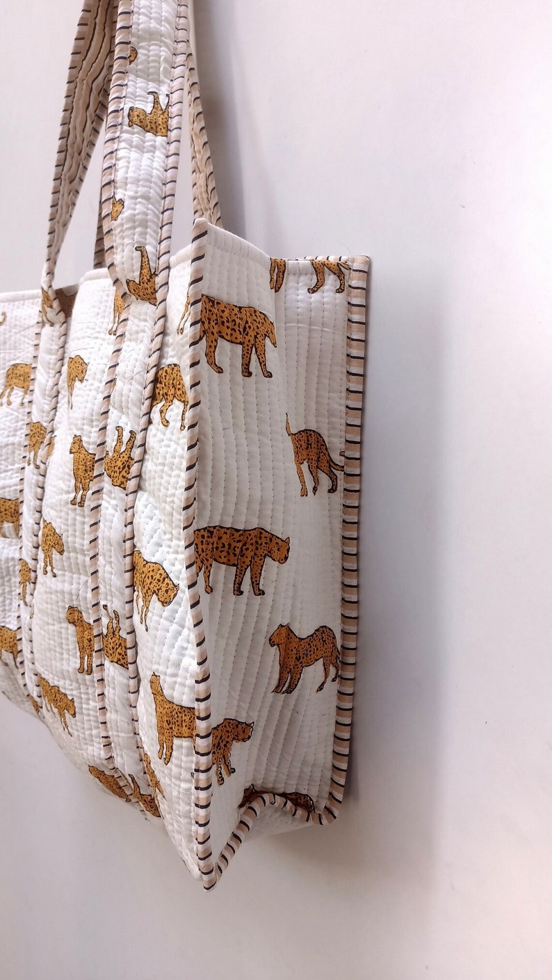 May include: White quilted tote bag featuring a repeating pattern of brown leopard illustrations. The bag has matching handles and trim with a striped design. The bag is made of a soft, quilted material.