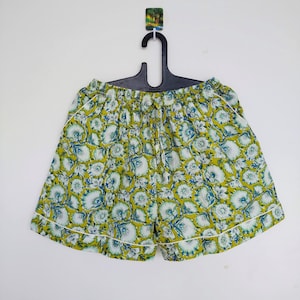 May include: Floral print shorts in shades of yellow, teal, and white, trimmed with white piping. The shorts are hanging on a black hanger against a white background. The fabric features a repeating pattern of stylized flowers.