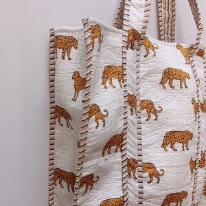 May include: White tote bag featuring a repeating pattern of orange and brown leopard print. The bag has matching handles and is trimmed with a striped border. The bag is made of a quilted fabric.