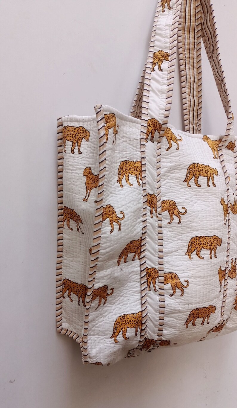 May include: White tote bag featuring a repeating pattern of orange and brown leopard print. The bag has matching handles and is trimmed with a striped border. The bag is made of a quilted fabric.