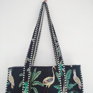 K&ouml;nnte beinhalten: Eine schwarze gesteppte Tragetasche mit wei&szlig;-schwarzem Besatz. Die Tasche hat einen tropischen Druck mit Palmen, Flamingos und V&ouml;geln. Die Tasche hat zwei lange Henkel.