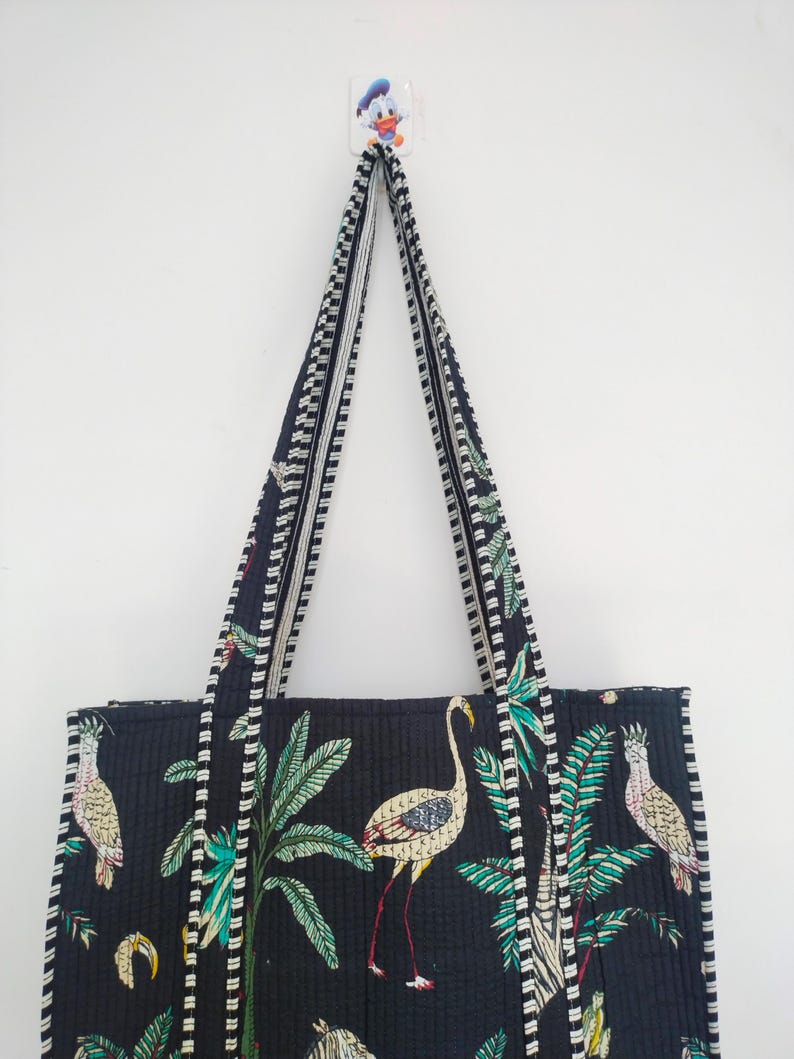 K&ouml;nnte beinhalten: Eine schwarze gesteppte Tragetasche mit wei&szlig;-schwarzem Besatz. Die Tasche hat einen tropischen Druck mit Palmen, Flamingos und V&ouml;geln. Die Tasche hat zwei lange Henkel.