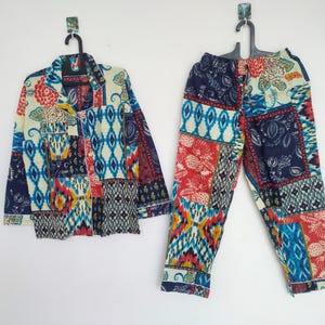 Peut inclure: Un ensemble de pyjama deux pièces avec un motif patchwork. L'ensemble comprend une chemise boutonnée et un pantalon assorti. Le tissu présente un mélange de motifs rouges, bleus, jaunes et blancs. Le pyjama est accroché à un mur blanc.
