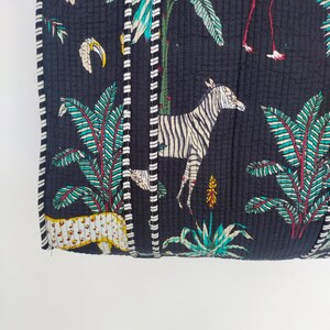 K&ouml;nnte beinhalten: Schwarzer gesteppter Stoff mit wei&szlig;em und schwarzem Besatz. Der Stoff zeigt ein sich wiederholendes Muster aus tropischen Pflanzen, Zebras, Tukanen und Flamingos.