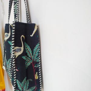 K&ouml;nnte beinhalten: Eine schwarze gesteppte Tragetasche mit wei&szlig;-schwarzem Besatz. Die Tasche hat einen floralen und Vogel-Druck in Gr&uuml;n-, Wei&szlig;- und Gelbt&ouml;nen.