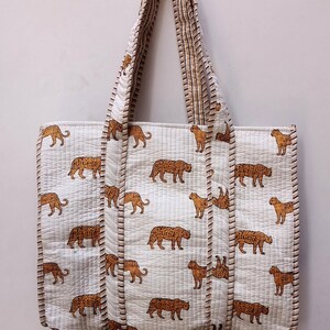 May include: White quilted tote bag featuring a repeating pattern of brown leopards. The bag has matching handles and trim. It is designed for carrying items. The bag is hanging against a white wall.