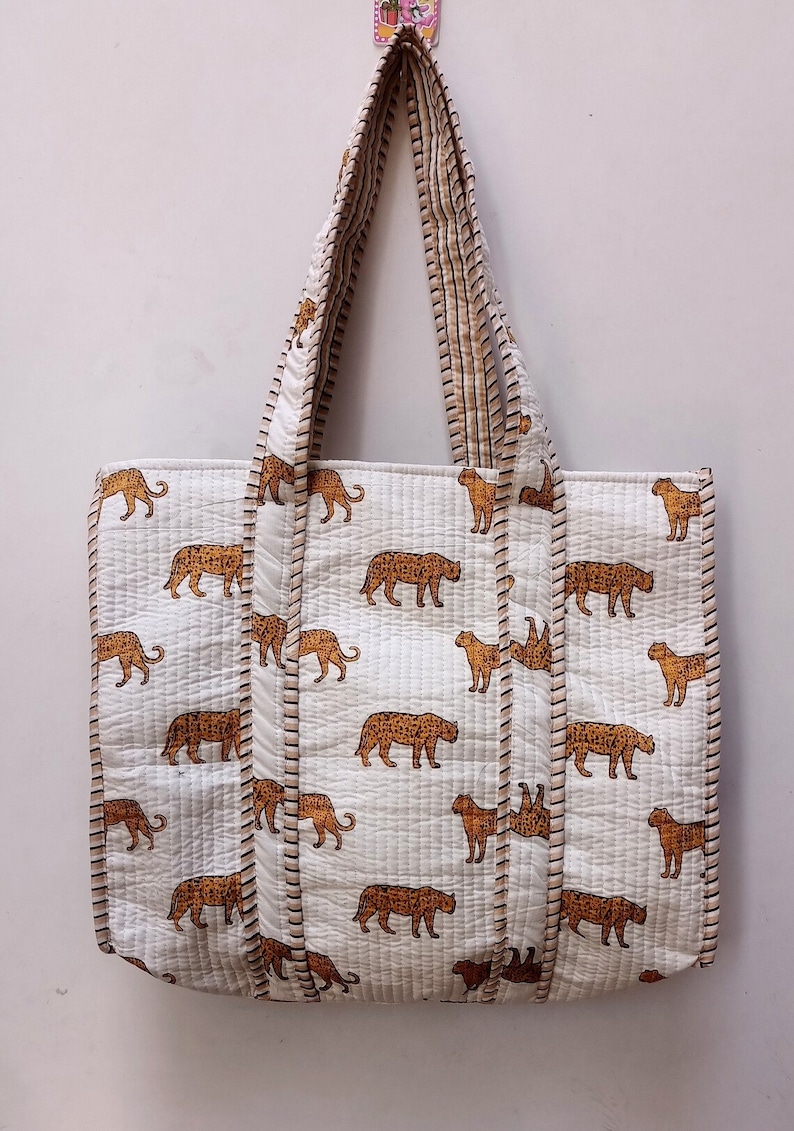 May include: White quilted tote bag featuring a repeating pattern of brown leopards. The bag has matching handles and trim. It is designed for carrying items. The bag is hanging against a white wall.