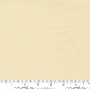 May include: A close-up of a piece of fabric in a pale yellow color. The fabric has a subtle texture and is measured with an inch ruler along the bottom edge. The brand name "moda" is printed on the ruler.