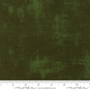 May include: Close-up of olive green fabric with a textured, distressed appearance. The fabric has a mottled effect with lighter green patches and subtle variations in tone. A ruler is at the bottom, with the word "moda" and the website address.