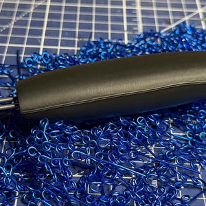 May include: A black, oval-shaped object with two silver metal rods extending from one end, resting on a bed of bright blue, wire-like decorative elements. The background is a blue grid.